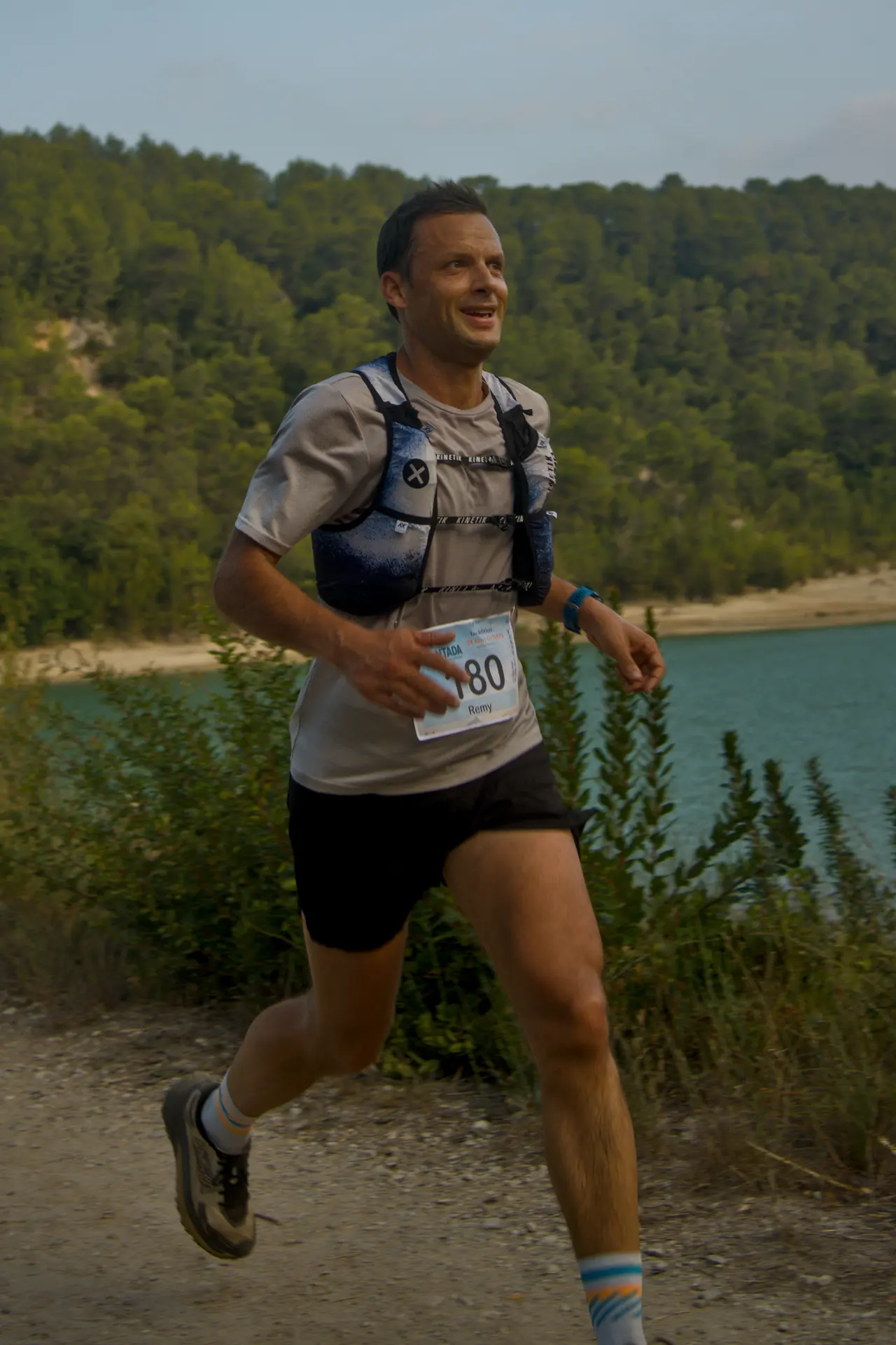 Un coureur souriant, dossard épinglé, longe un lac turquoise bordé de pins.