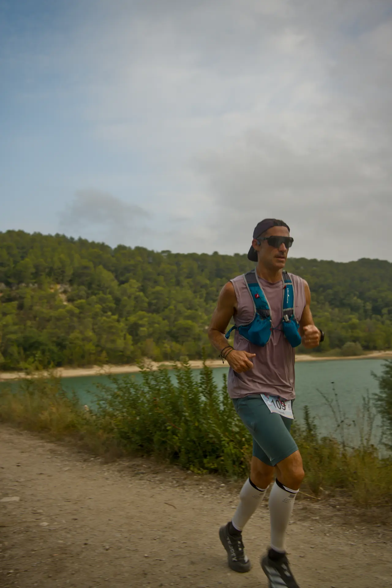 Un coureur en lunettes de soleil et gilet d’hydratation longe un lac.